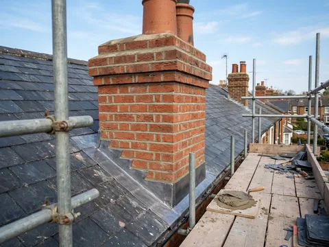 Repointed red brick chimney stack with new lead flashing where it meets the slate roof, seen from the scaffold